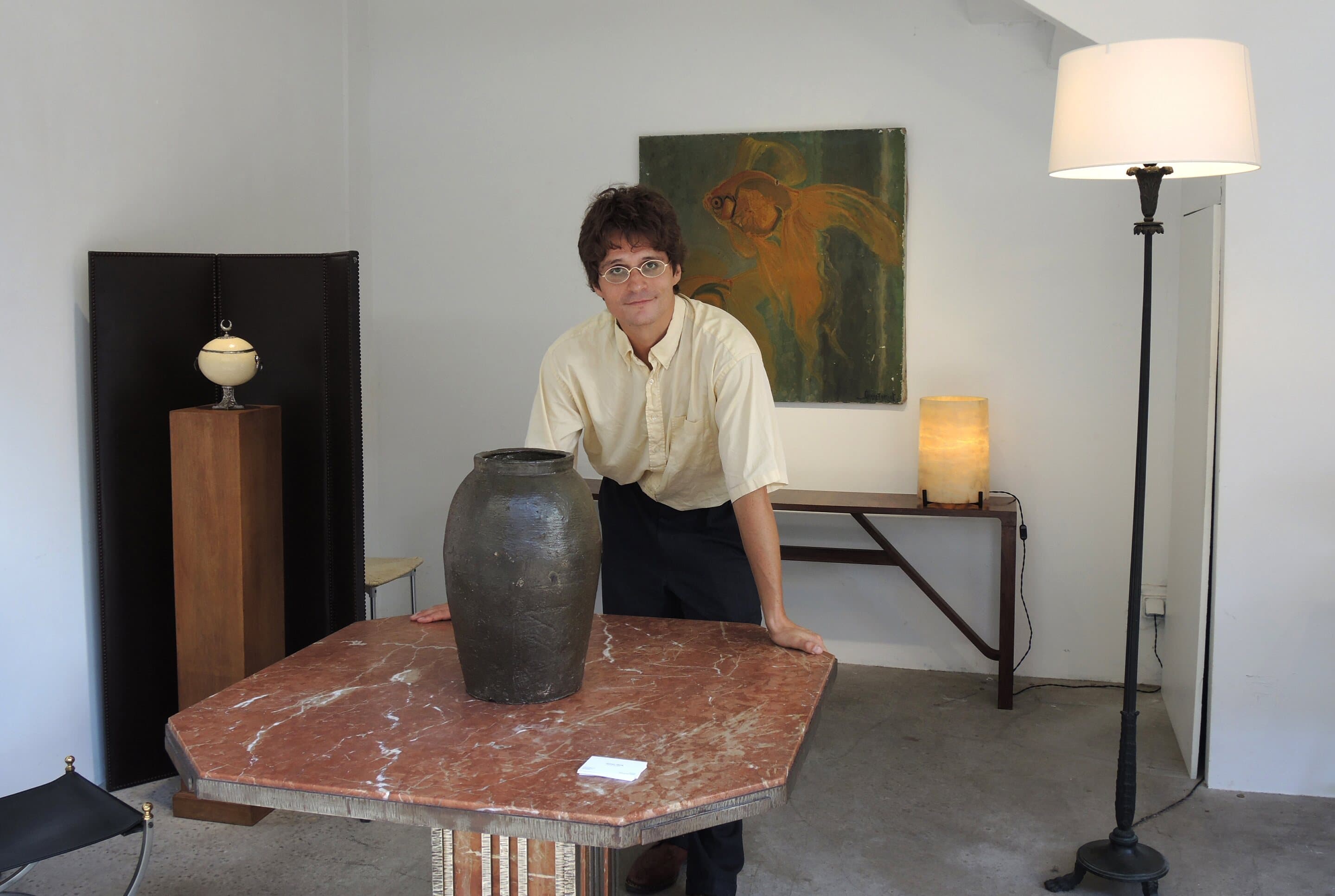 James Darle at his stand, leaning on an octagonal red marble table beside a stoneware jar