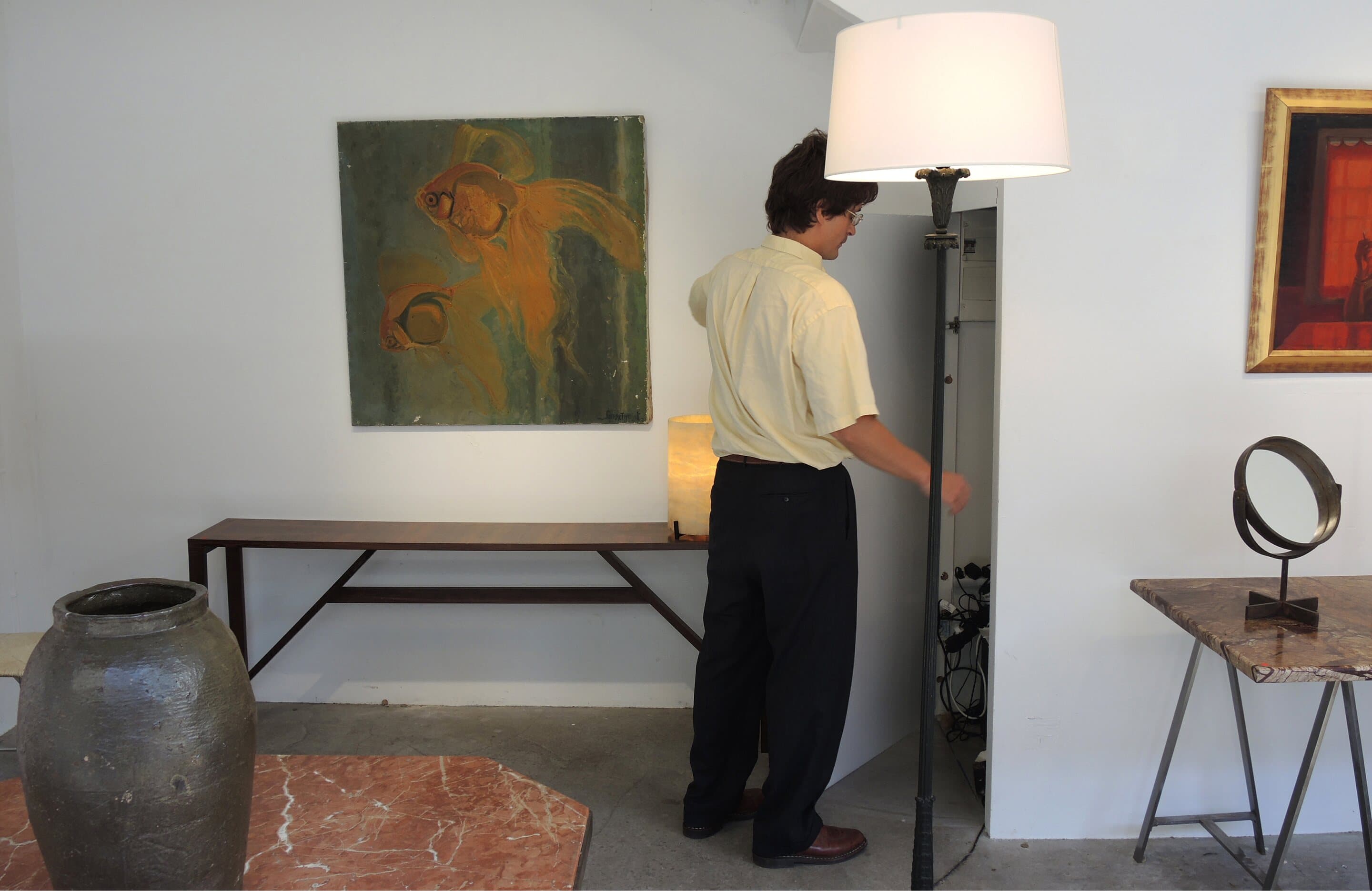 James Darle at his stand, opening a cupboard beside a floor lamp, a painting of goldfish on the wall